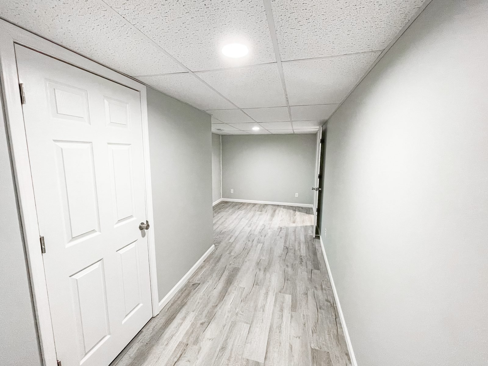 Basement hallway in Cookeville with a white six-panel door, drop ceiling, recessed light and vinyl plank flooring after an H&H Construction remodel
